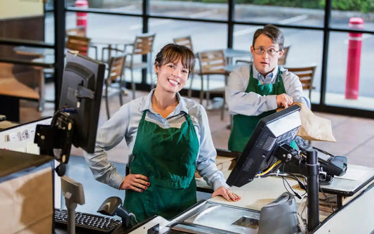 A smiling female cashier in a green apron standing at a checkout counter in a retail or food service setting, with a male colleague working in the background