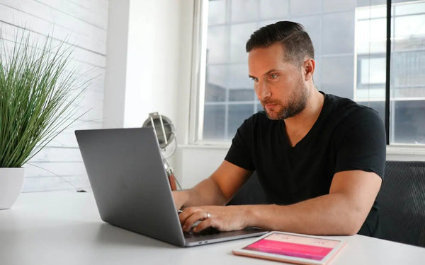 A man in a black t-shirt working on a laptop with a tablet next to it on a white desk in a bright office environment