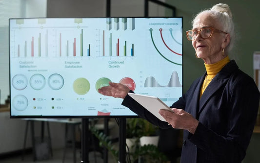 An older woman wearing glasses and a dark blazer holding a tablet while giving a presentation in front of a large screen displaying charts and data about business metrics