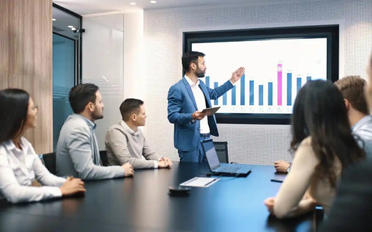 A man wearing a blue suit and holding a tablet while presenting business charts on a large screen to colleagues seated around a conference table in an office