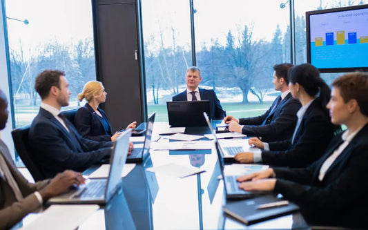 A group of professionals in suits sitting around a large glass conference table in a bright corporate meeting room with laptops open, while a man leads the discussion and a presentation screen is visible in the background