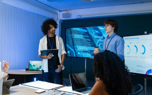 A group of professionals collaborating in a modern office during a meeting, with two men standing and presenting strategies in front of large screens displaying digital graphics and business data charts