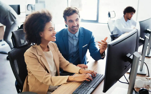 A smiling woman typing on a computer keyboard with a male colleague looking over her shoulder pointing at the screen in a bright office environment