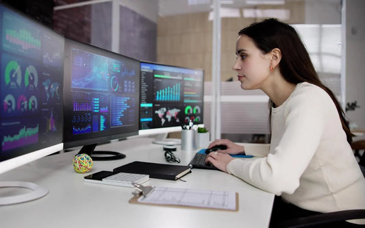 A professional woman working at a desk with multiple computer monitors displaying data analytics and charts in an office setting