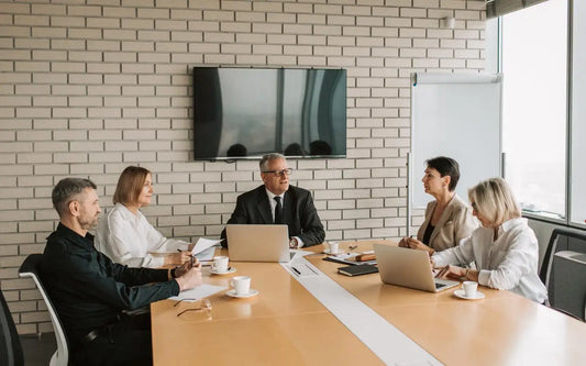 A group of professionals in a boardroom meeting around a long wooden table, with laptops, documents, and coffee cups set up against a brick wall background