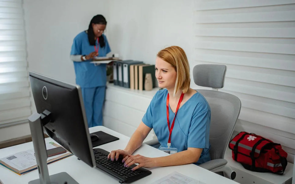 A healthcare professional working on a computer at a desk in an office setting, with another medical staff member standing in the background and a first aid bag on the floor