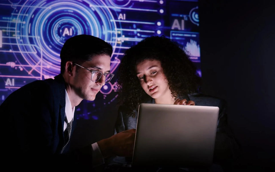 A man wearing glasses and a woman working together on a laptop in a dimly lit room with a blue illuminated digital graphic background displaying AI symbols
