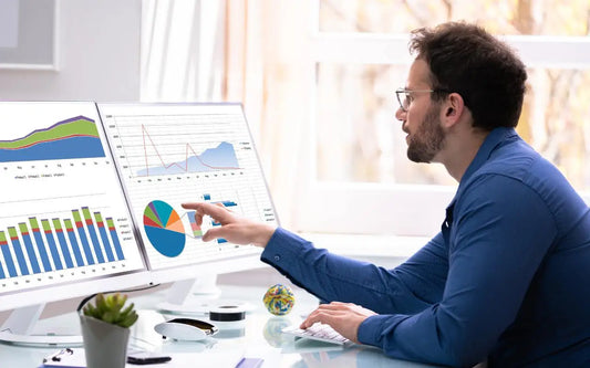 A man wearing glasses and a blue shirt sitting at a desk with dual monitors displaying financial charts and graphs, pointing at a pie chart