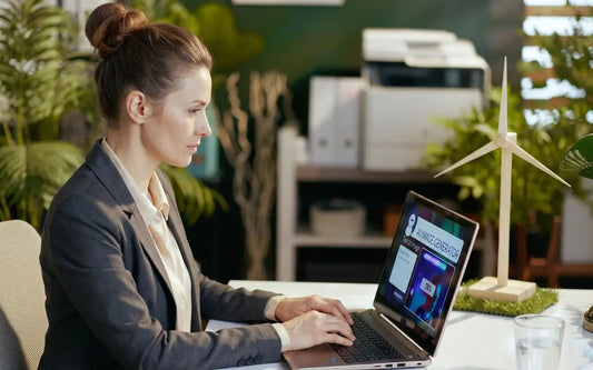 A professional woman with her hair in a bun working on a laptop at a desk with a small wind turbine model, with an AI image generator interface visible on the screen