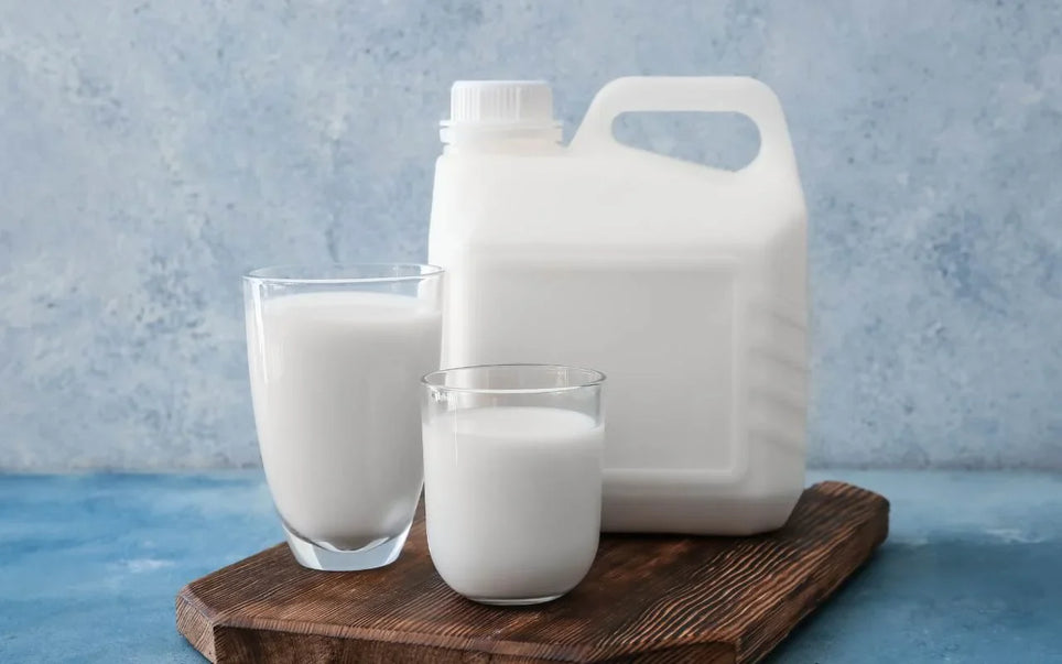 A large plastic container and two glasses filled with white milk placed on a wooden board against a textured blue background