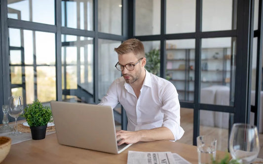 A man wearing glasses and a white shirt working on a laptop at a wooden table in a room with large glass windows and black frames