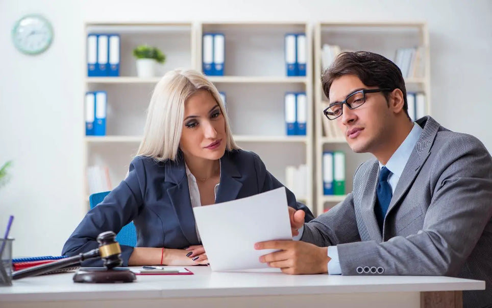 A professional man wearing glasses and a suit reviewing legal documents with a female colleague next to a desk with a gavel in an office