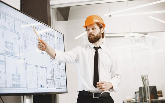 An architect wearing an orange hard hat, white shirt, and tie pointing at a floor plan blueprint displayed on a large screen during a presentation