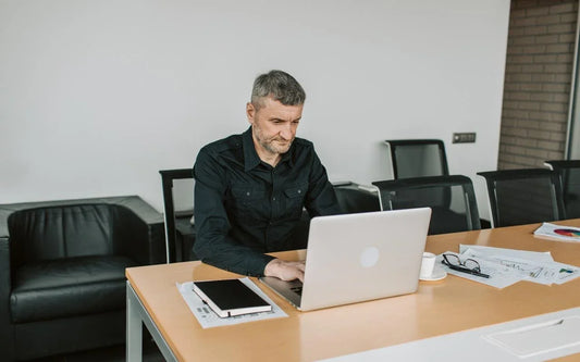 A middle-aged man working on a laptop at a long wooden conference table in an office setting, with a notebook, glasses, and papers nearby.
