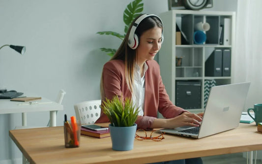 A young woman wearing headphones and a pink blazer working on a laptop at a wooden desk in a home office setup with a bookshelf in the background