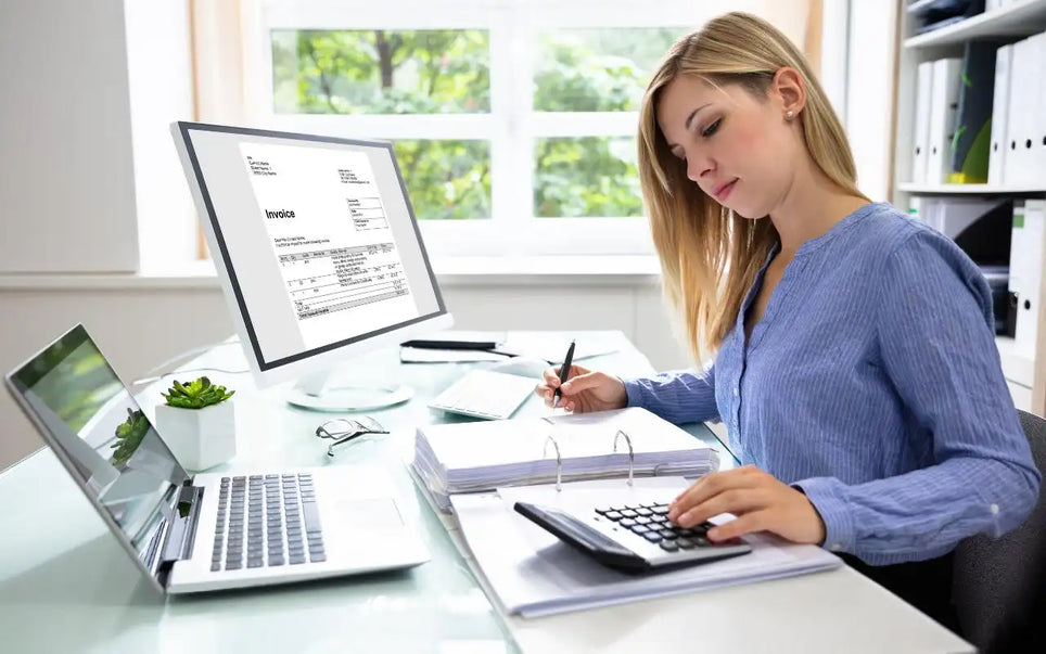 A professional woman working at a desk with a laptop and a desktop monitor displaying an invoice, using a calculator and reviewing documents in an office setting
