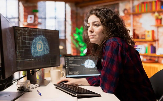 A woman with curly dark hair wearing a red and black plaid shirt sitting at a desk with multiple monitors and a laptop displaying code and digital graphics