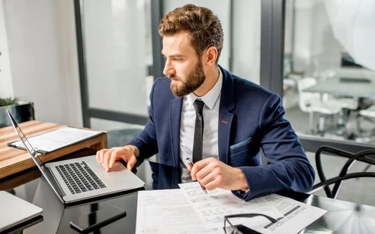 A professional bearded man wearing a suit and tie working on a laptop at a sleek glass desk with documents and papers spread out around him
