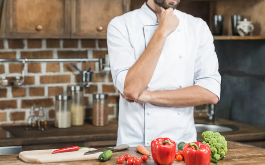 Thoughtful chef standing behind a kitchen counter with fresh vegetables and food-preparation equipment.